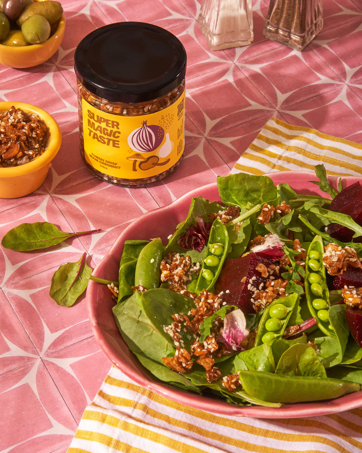 A spinach and beet salad topped with a scattering of Super Magic Taste Yummy Crisp, the jar on a pink cloth behind it.