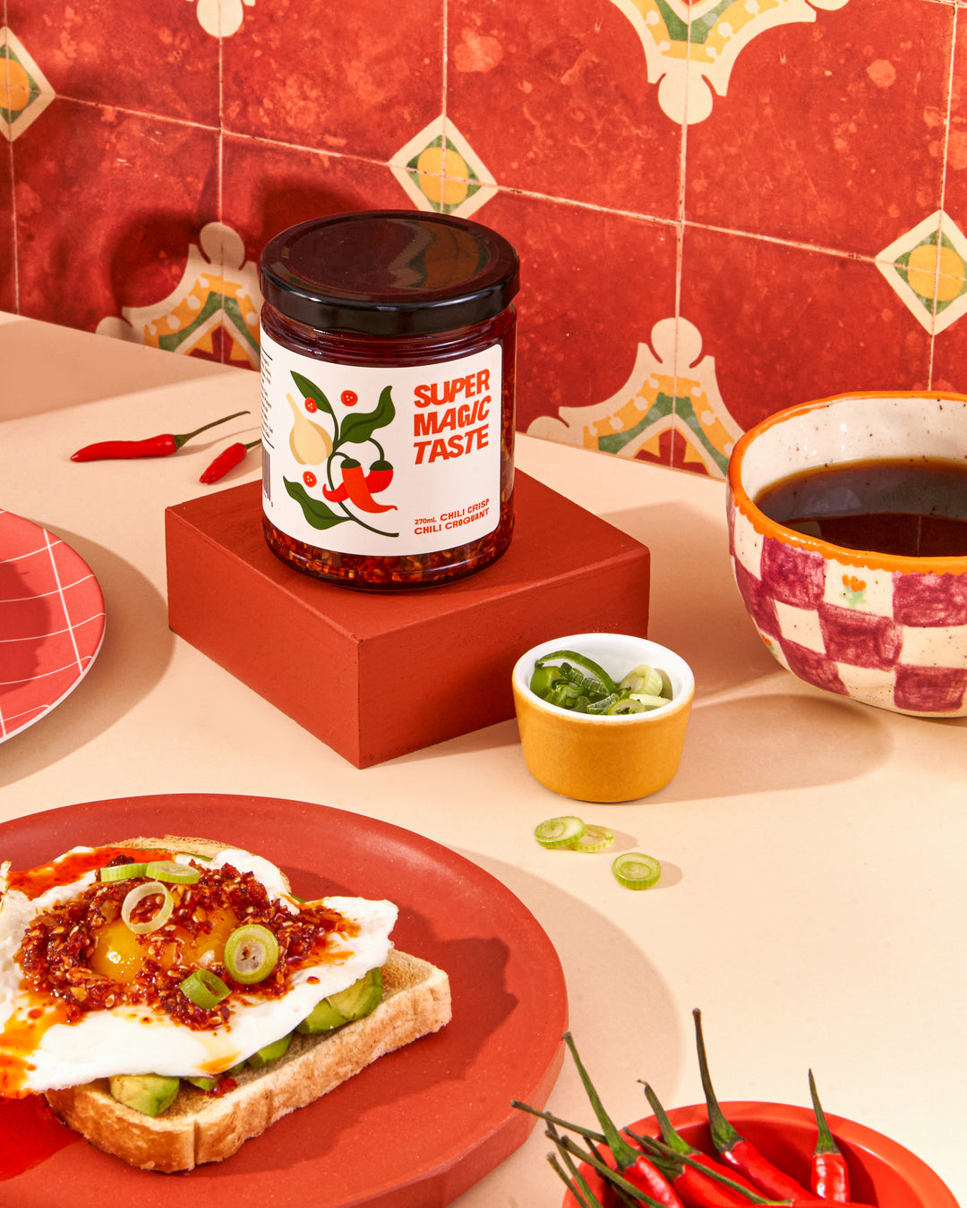 Super Magic Taste Chili Crisp spooned over a fried egg on avocado toast, with the open jar and a cup of coffee behind.