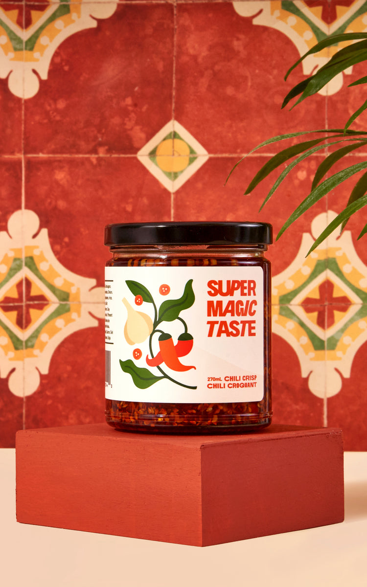 A jar of Super Magic Taste Chili Crisp on a terracotta plinth against red and green patterned Spanish tile.